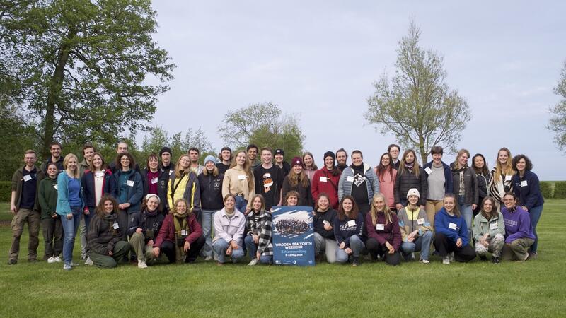 Group photo of 40 young people holding a "wadden sea youth weekend" sign, with blue sky, grass and some trees behind the group. P. Ertzinger.